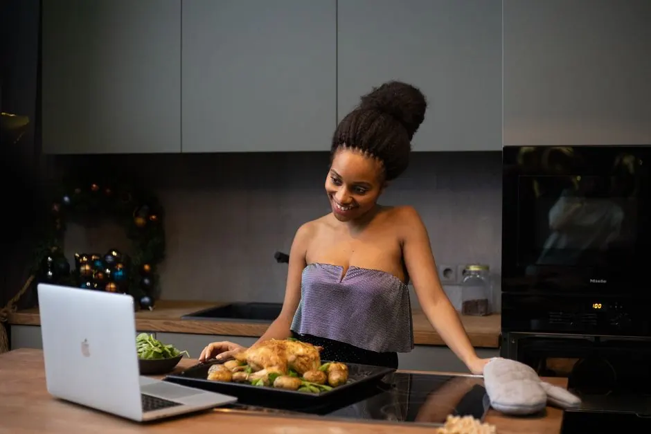 African American woman in kitchen smiling at a laptop with a tray of roast chicken and vegetables.
